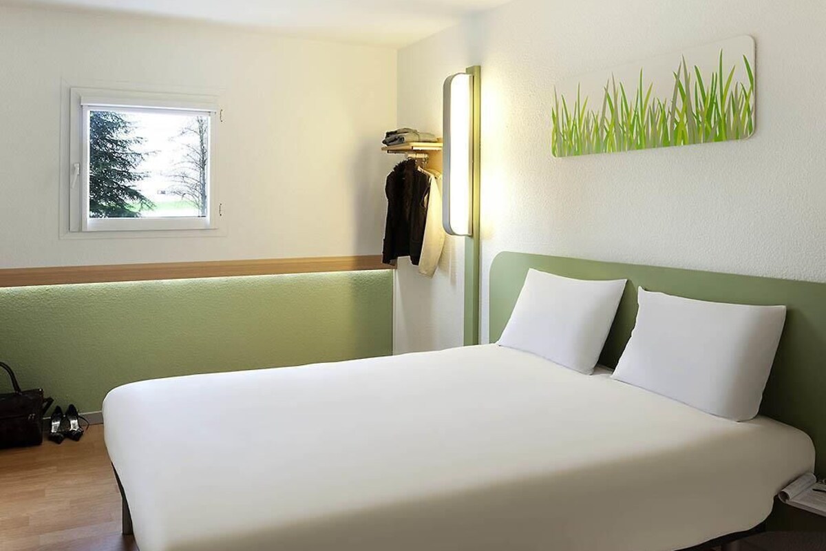 Une chambre d’hôtel avec un lit, une fenêtre, une lampe fixée au mur et une œuvre d’art décorative.