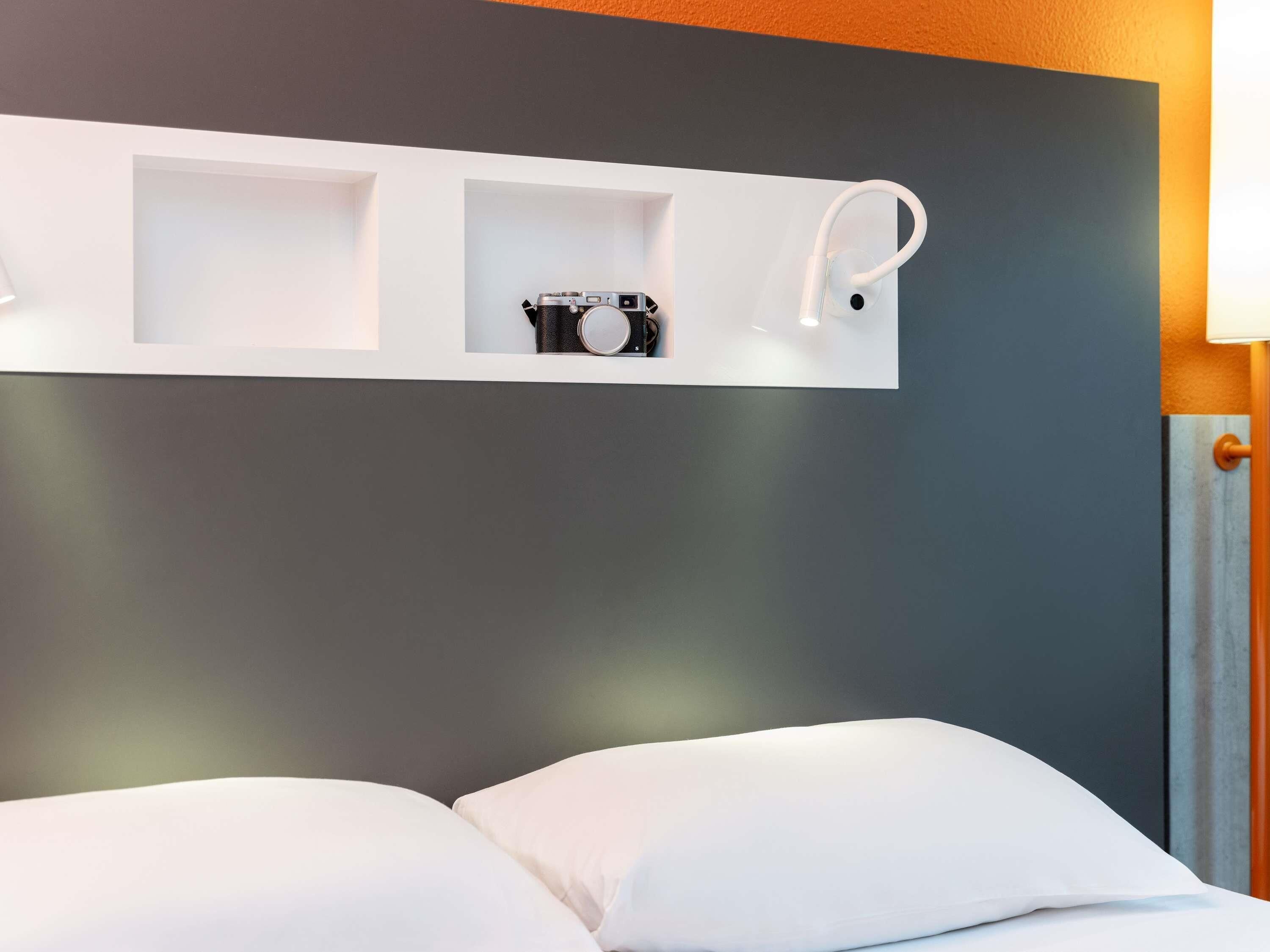 Ein Hotelzimmer mit einem Bett, einer weißen Ablage mit Kamera und Lampe sowie einer grauen Wand.