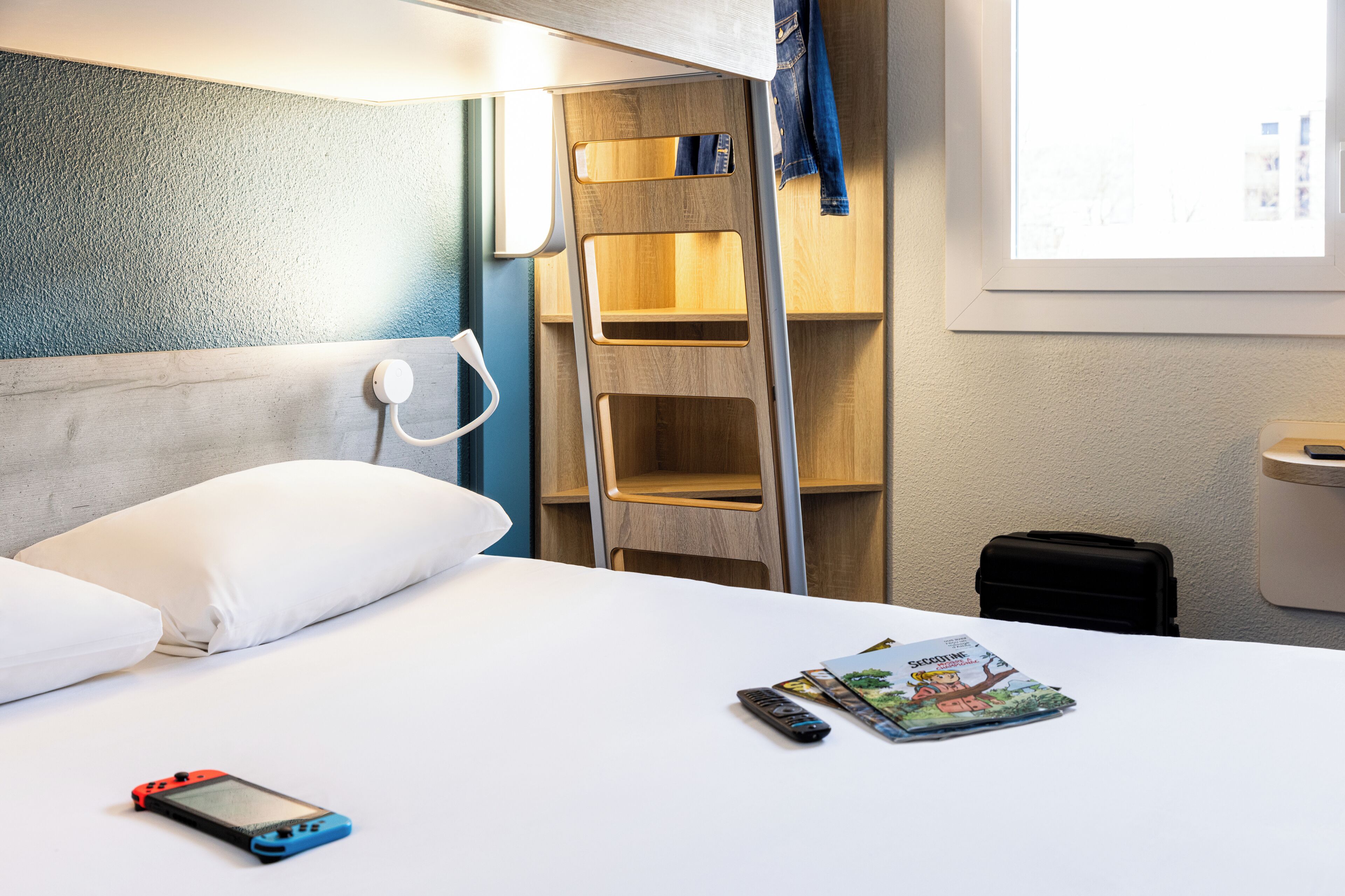 A hotel room with a bed, a ladder, a suitcase, and a magazine on the bed.