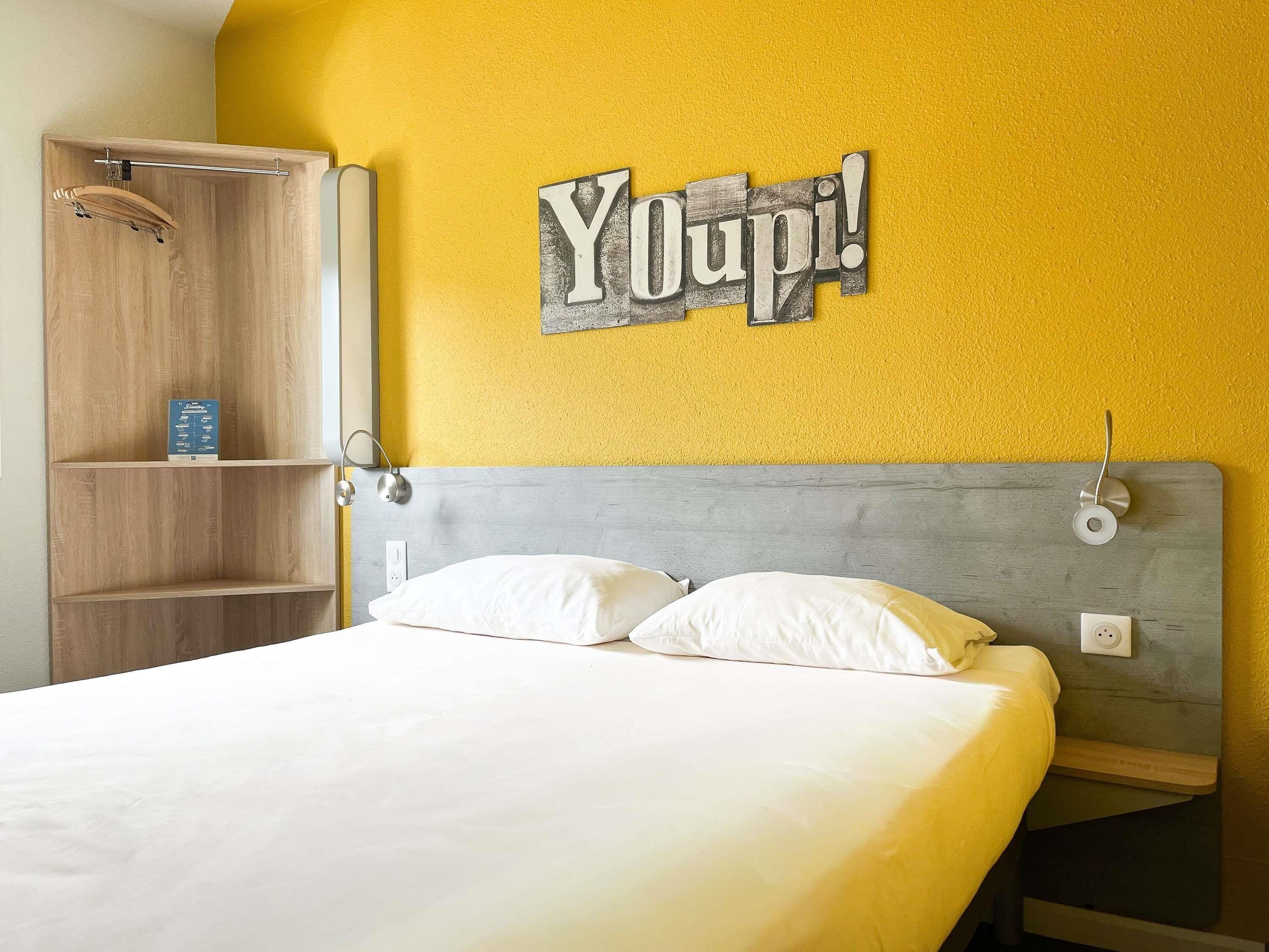 A bed with white bedding, a wooden wardrobe, and a wall-mounted sign that reads 'Youpi!'