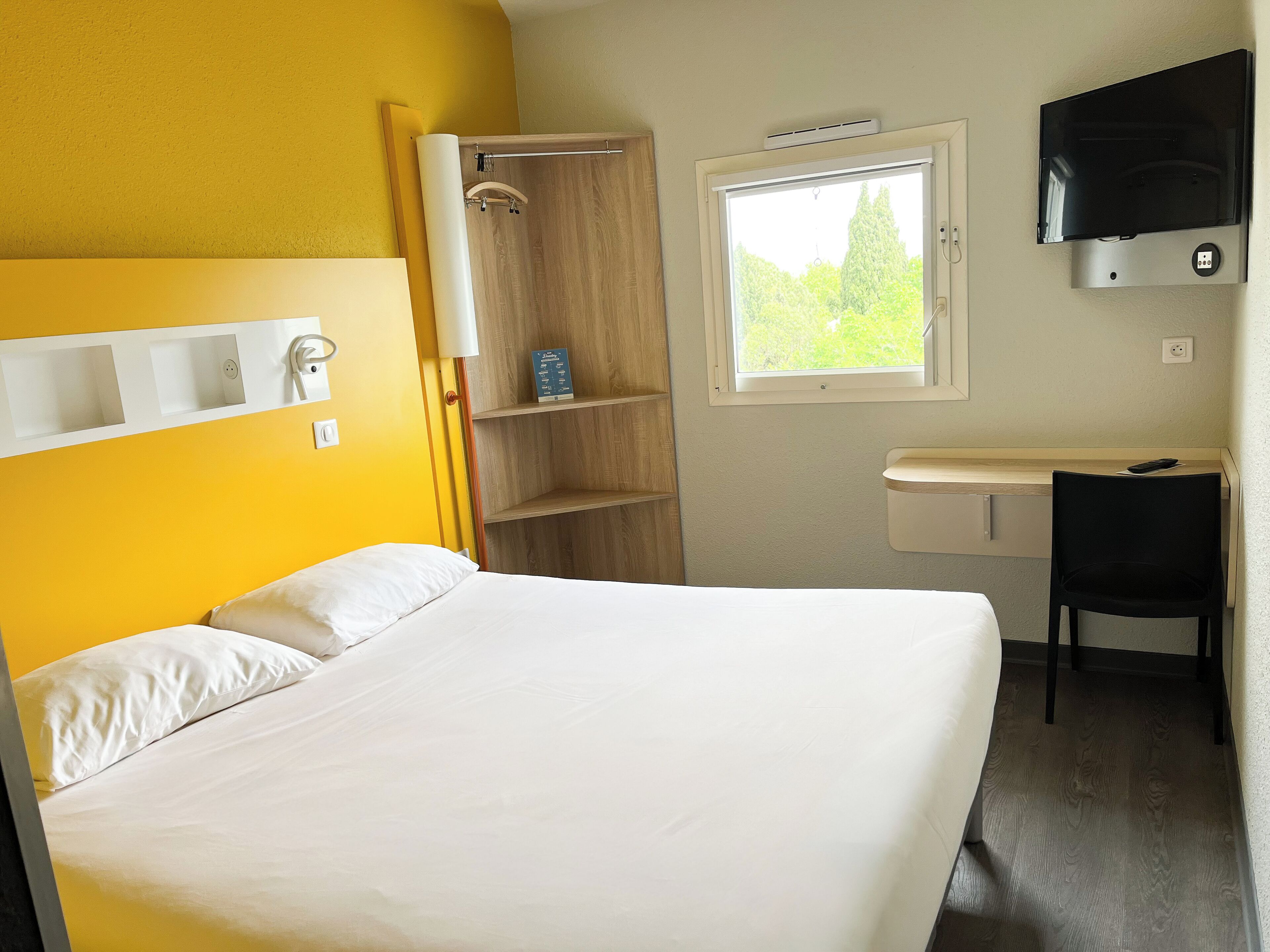Quarto de hotel com cama, escrivaninha, cadeira, televisão, janela e chuveiro.