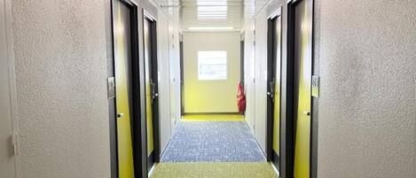 A hotel hallway with doors on both sides, a patterned carpet, and a window at the end.
