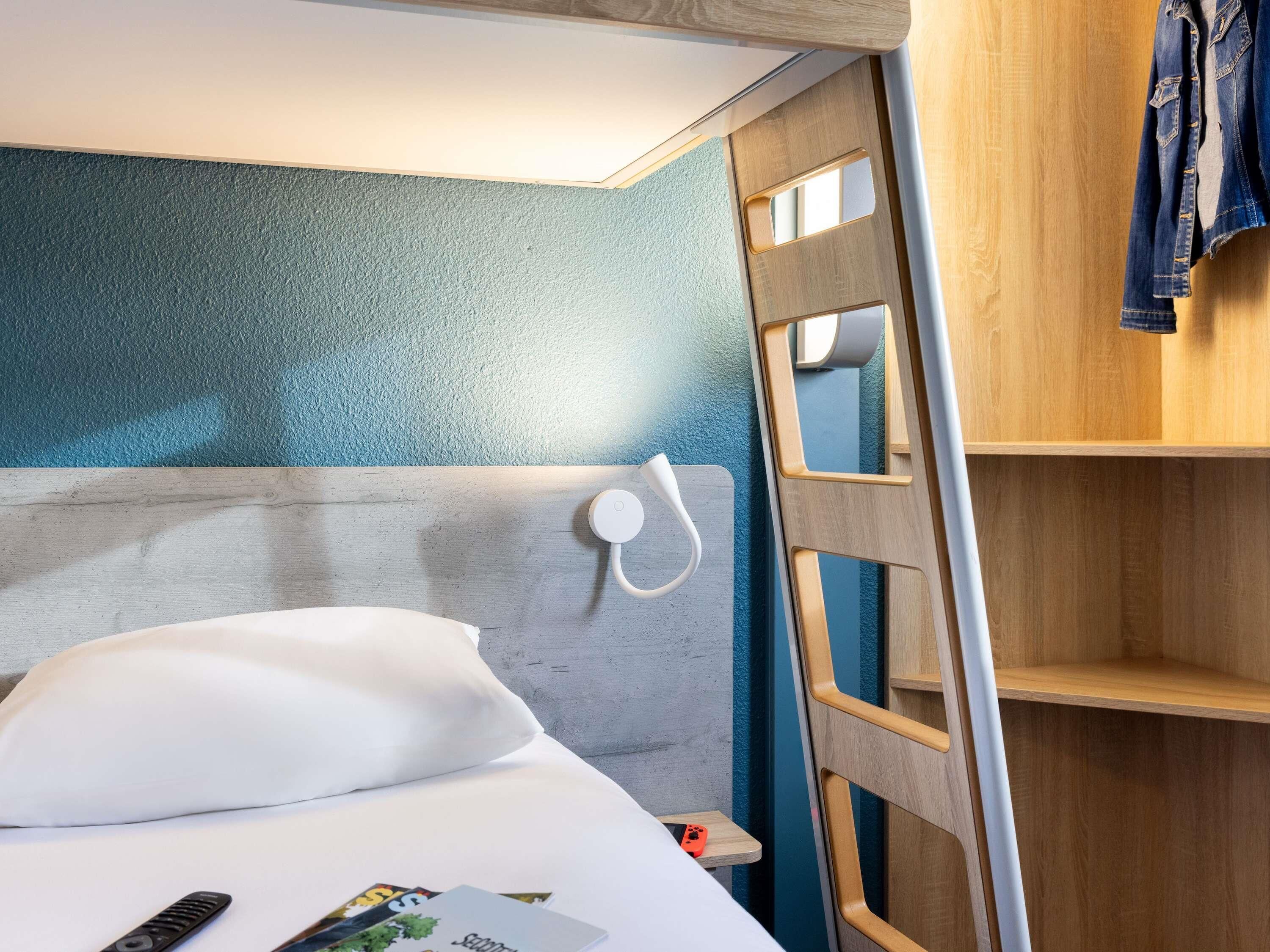 Un lit avec un oreiller blanc, une télécommande et un magazine dessus. Une échelle en bois est fixée à un mur, et une étagère en bois porte une veste.