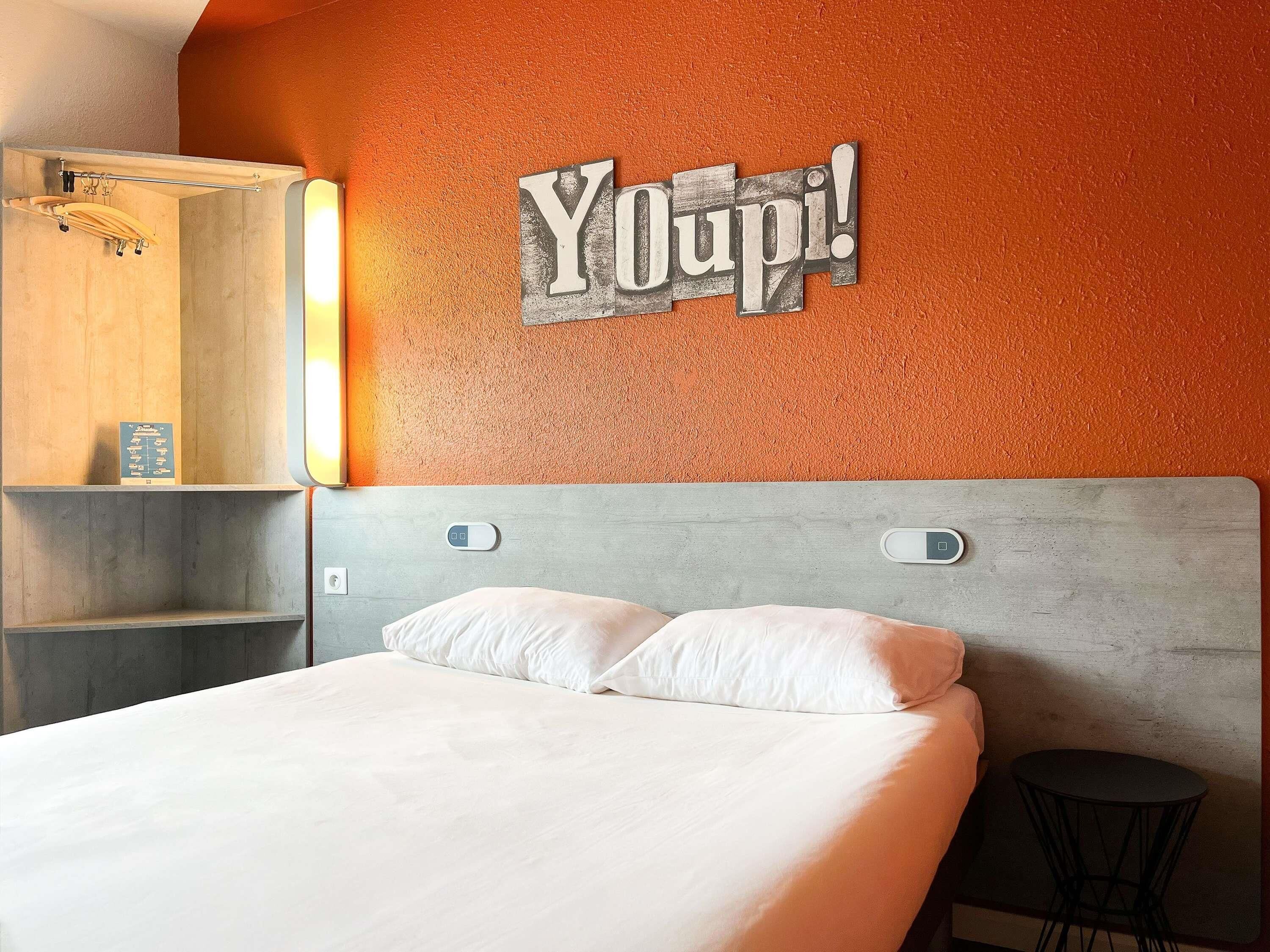 Une chambre d’hôtel avec un lit, une étagère en bois, un miroir et un panneau mural sur lequel on peut lire : « Youpi ! ».