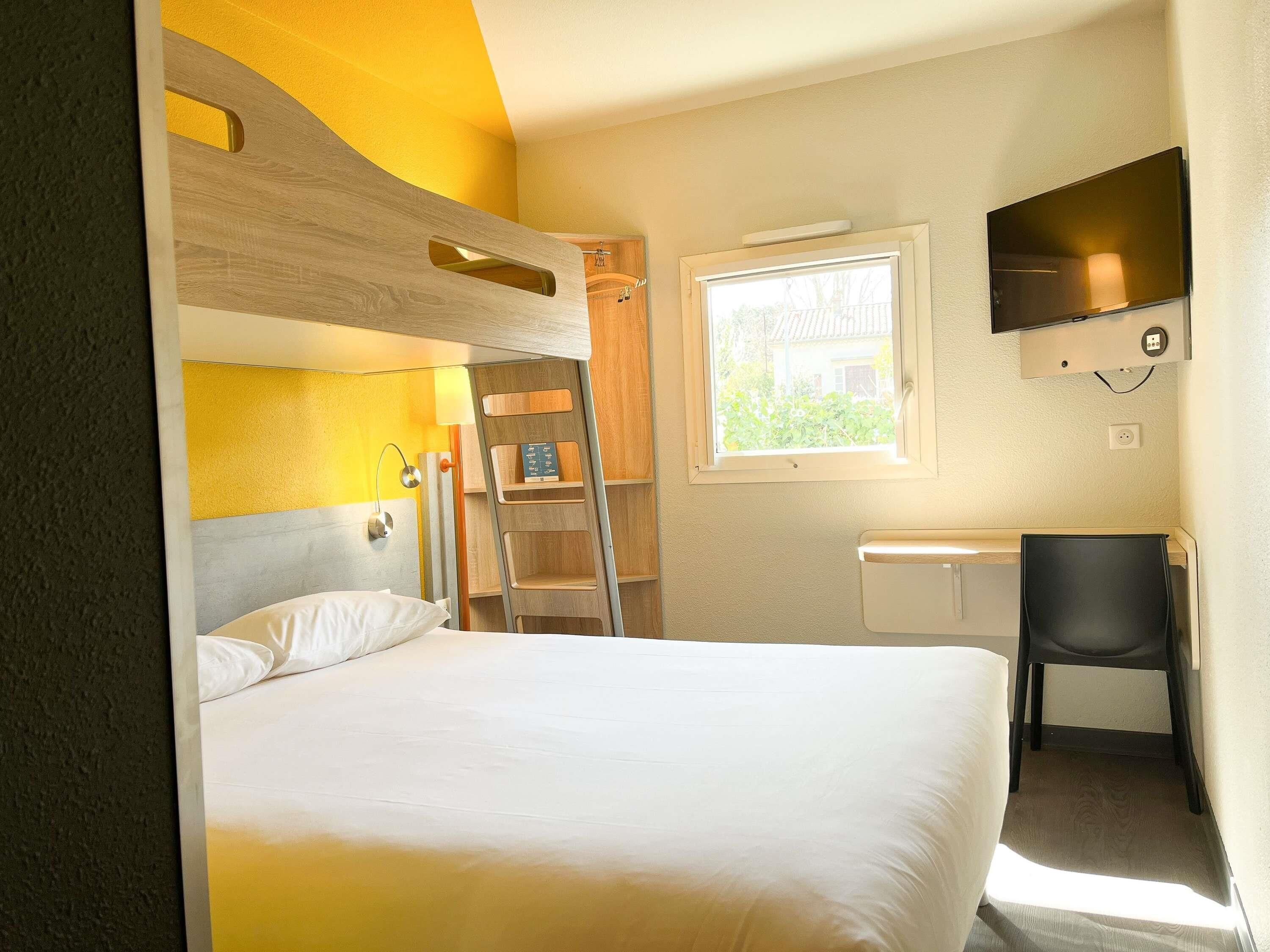 Ein Hotelzimmer mit Etagenbett, Schreibtisch und Stuhl, einem an der Wand montierten Fernseher und einem Fenster mit Blick auf Grünflächen.