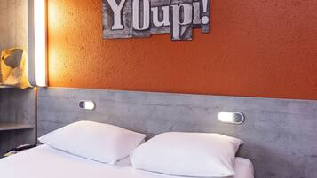 Quarto de hotel com cama, almofadas brancas, cabeceira e parede com o texto "Youpi!".