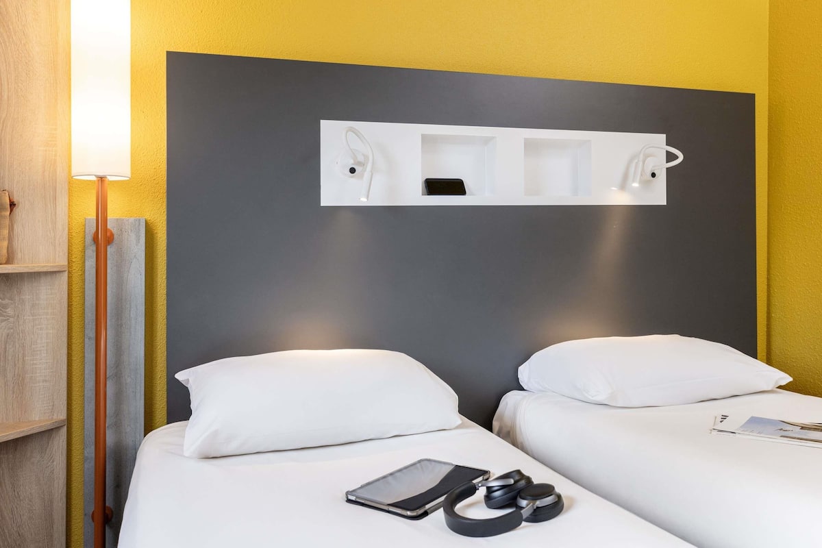 Habitación de hotel con dos camas, una mesita de noche, una lámpara y una estantería sobre las camas.