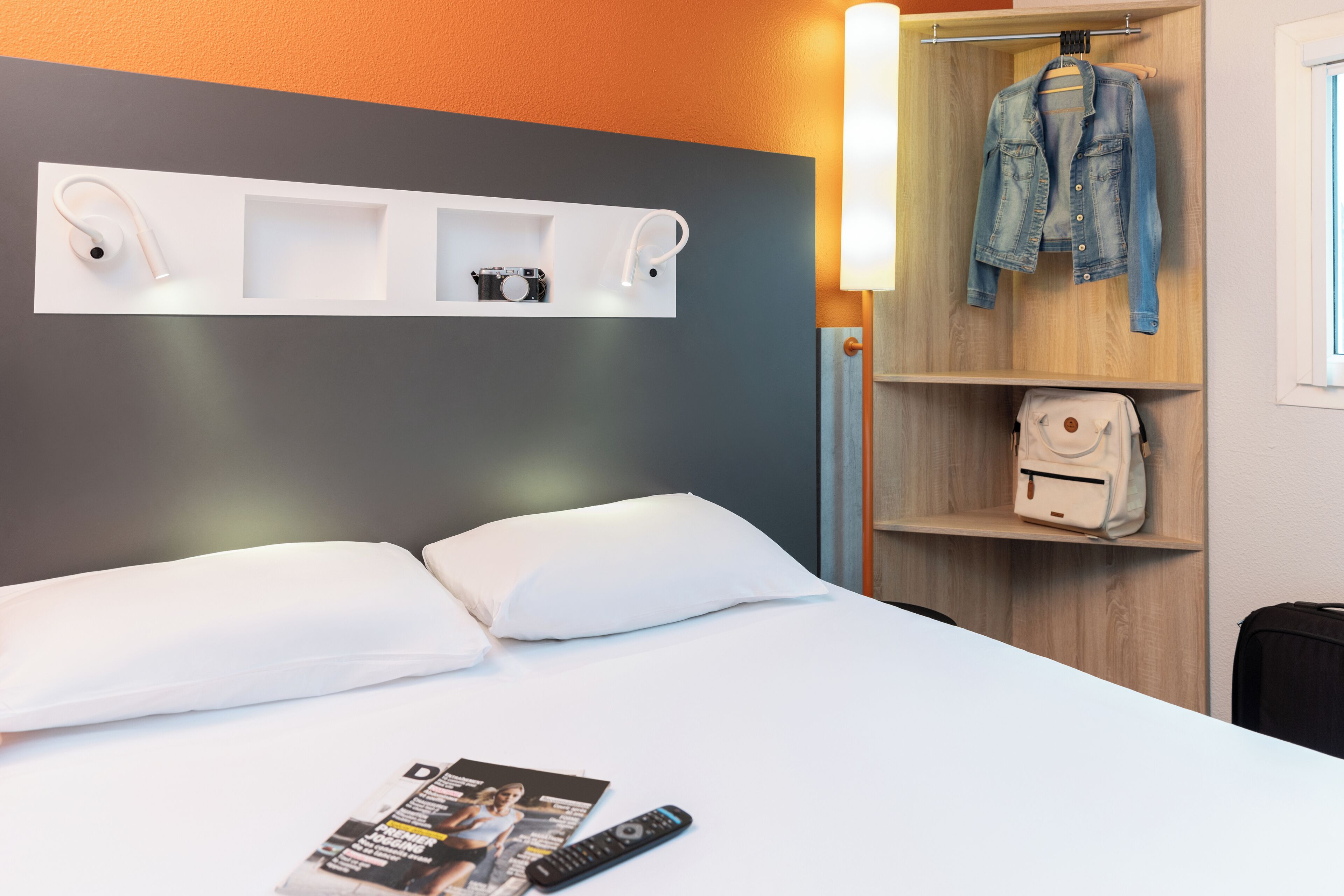 Habitación de hotel con cama, almohadas blancas, una revista, un control remoto, una cámara y una chaqueta vaquera colgada en un perchero.