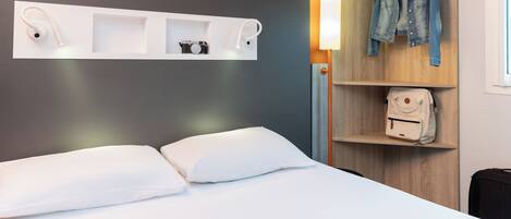 Habitación de hotel con cama, almohadas blancas, una revista, un control remoto, una cámara y una chaqueta vaquera colgada en un perchero.