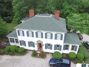 A two-story house with a grey roof, white walls, and blue shutters. There are two chimneys, a front door, and a driveway with two cars parked.