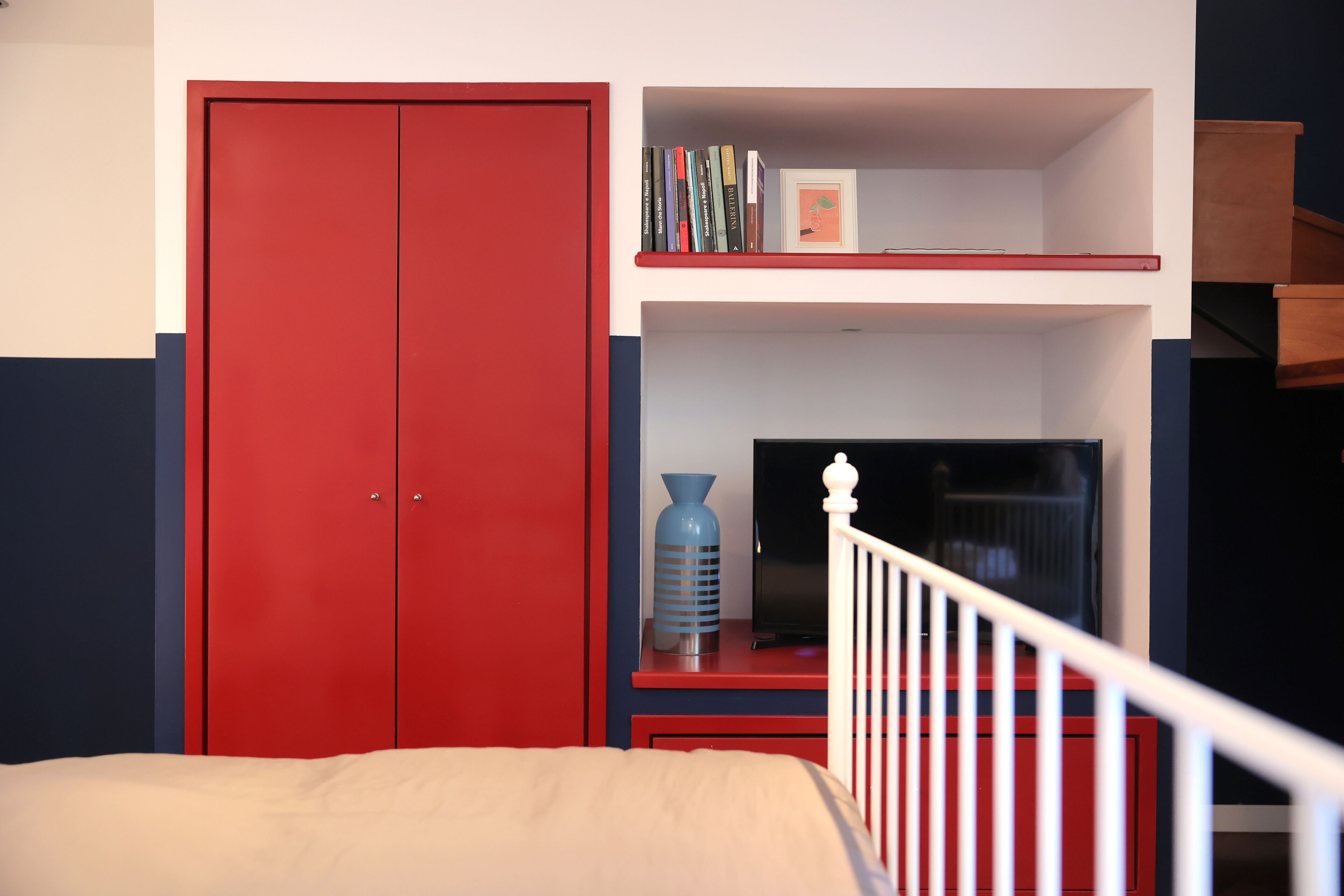 Un dormitorio con una cama, un armario rojo, una estantería con libros y un jarrón, y un televisor sobre un mueble.