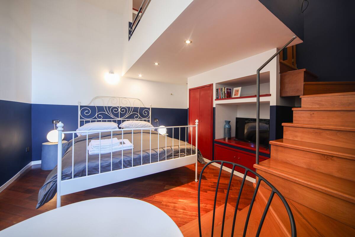 A bedroom with a bed, bedside table, chair, and a staircase.