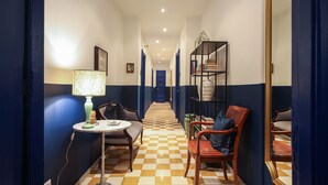 A hotel hallway with a checkered floor, a round table with a lamp, a sofa, and a chair. There is a bookshelf with books and a mirror on the wall. - Salotto Borbonico (Naples)