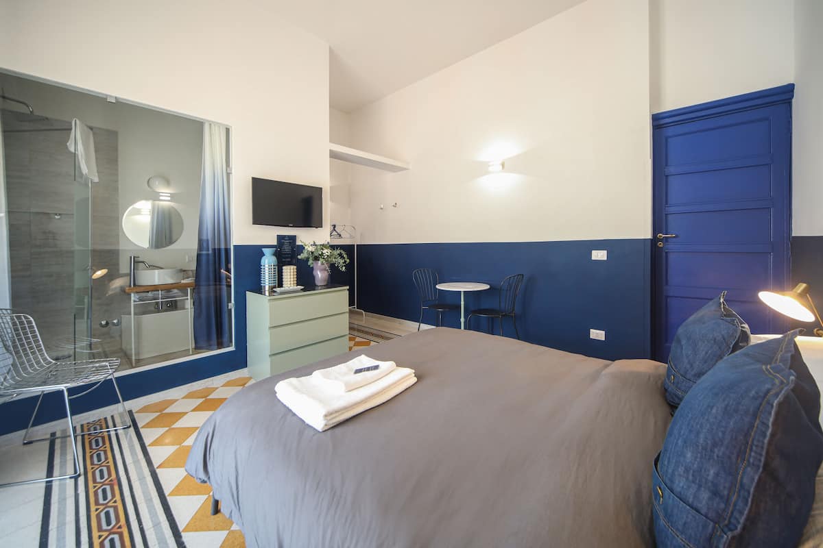 A modern hotel room with a large bed, a glass shower, a small desk with a chair, and a TV.