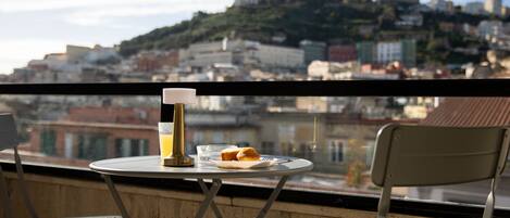 Ein Balkon mit Frühstückstisch, einem Glas Orangensaft und einem Gebäck. Im Hintergrund ist eine Stadtlandschaft mit Gebäuden und einem Hügel zu sehen.