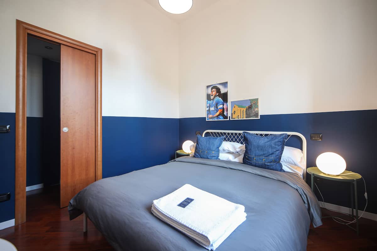 A bedroom with a bed, a nightstand with a lamp, a door, and wall art.