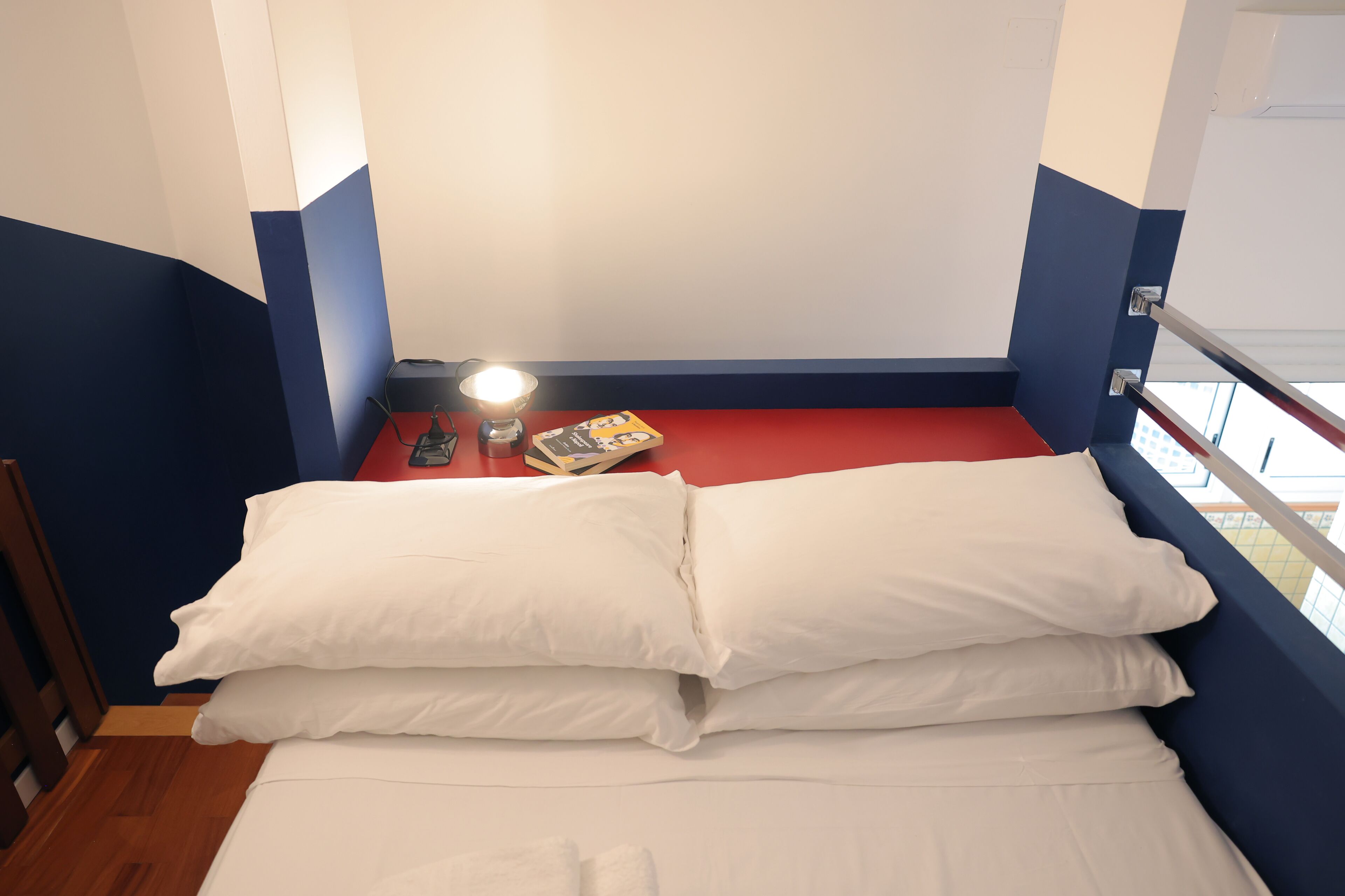 A small, compact room with a bed, a nightstand, a lamp, and a window.