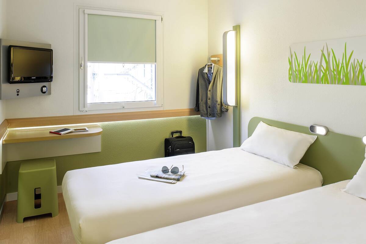 A hotel room with a bed, a desk with a computer, a window, and a wall-mounted TV.