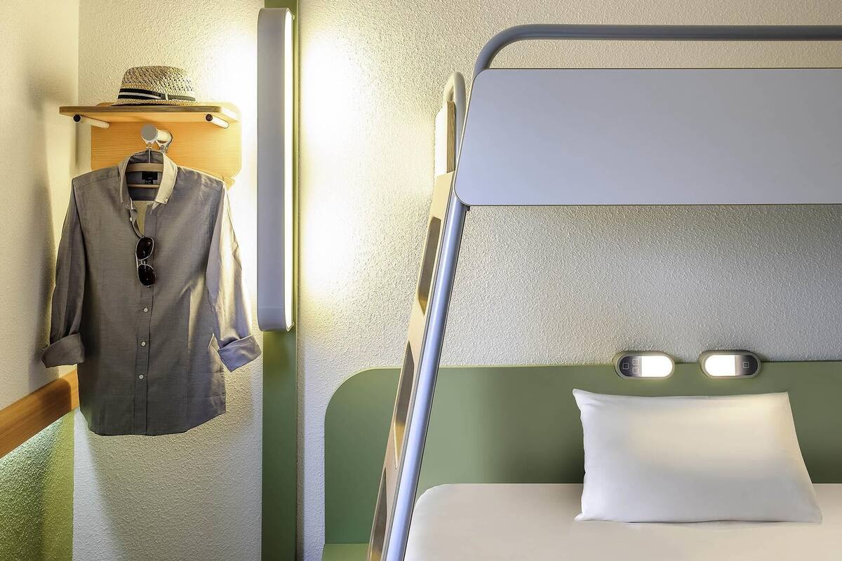 A bunk bed with a green headboard and a white pillow, a wooden hanger with a hat and a gray shirt, and a wall-mounted light fixture.