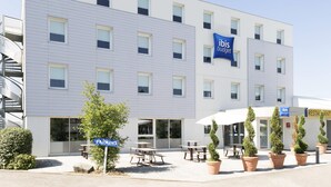 A modern building with a flat facade, multiple windows, and a sign for 'ibis budget'. There are outdoor tables and potted plants in front. - ibis Budget Lyon Eurexpo Chassieu (Chassieu)