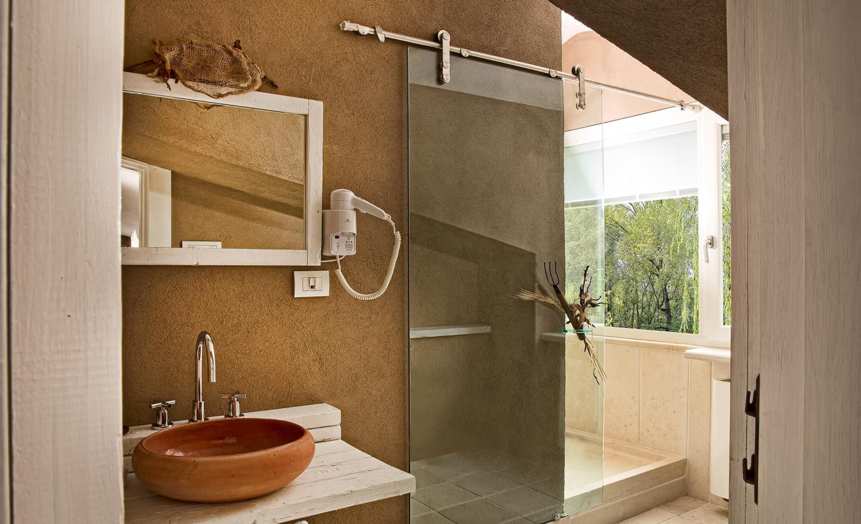 A bathroom with a large mirror, a round sink, a shower with a glass door, and a view of greenery outside.