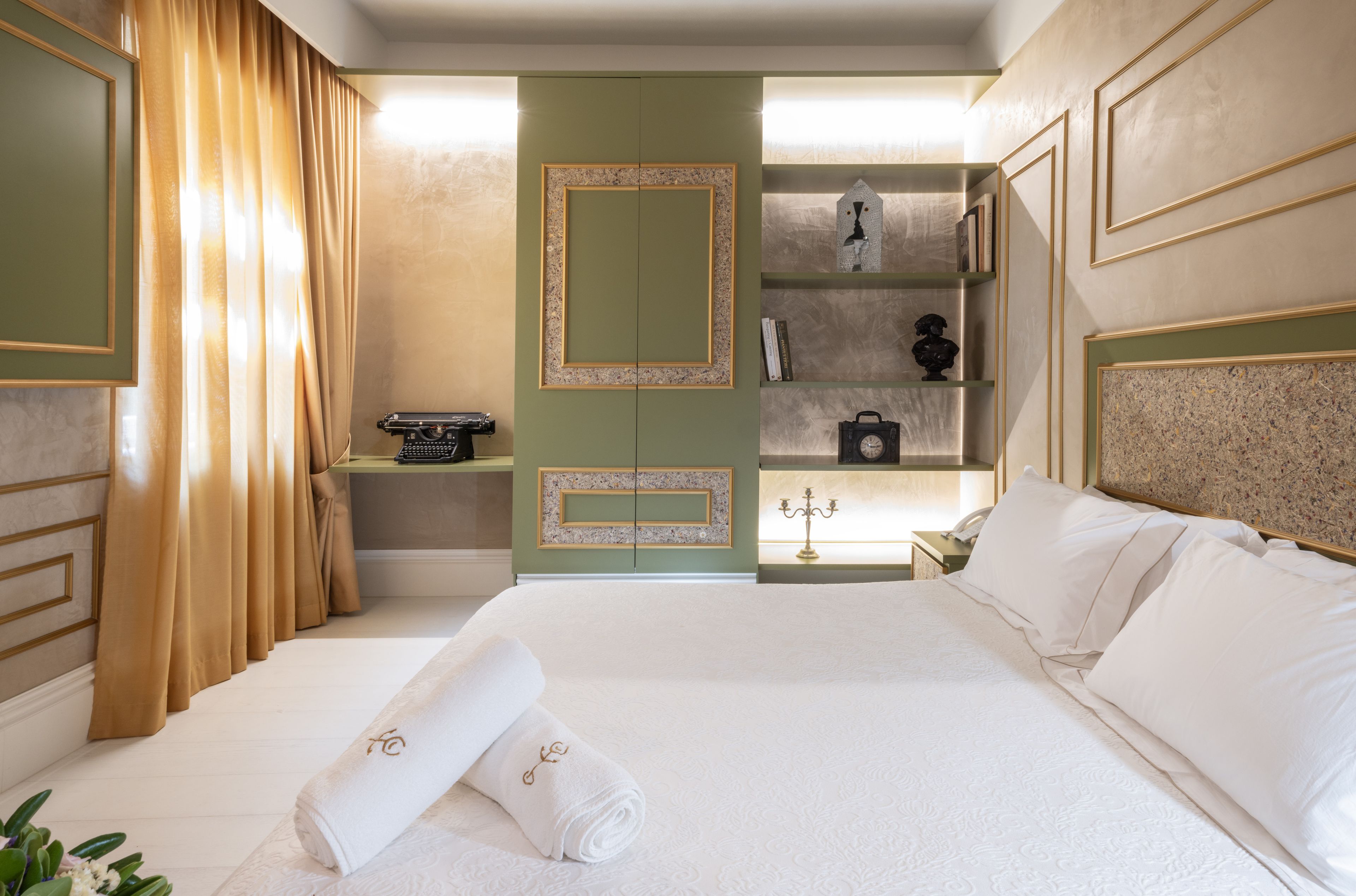 Une chambre d’hôtel avec un grand lit, un bureau avec un téléphone, une étagère avec des livres et un appareil photo, et une vue sur la ville par la fenêtre.