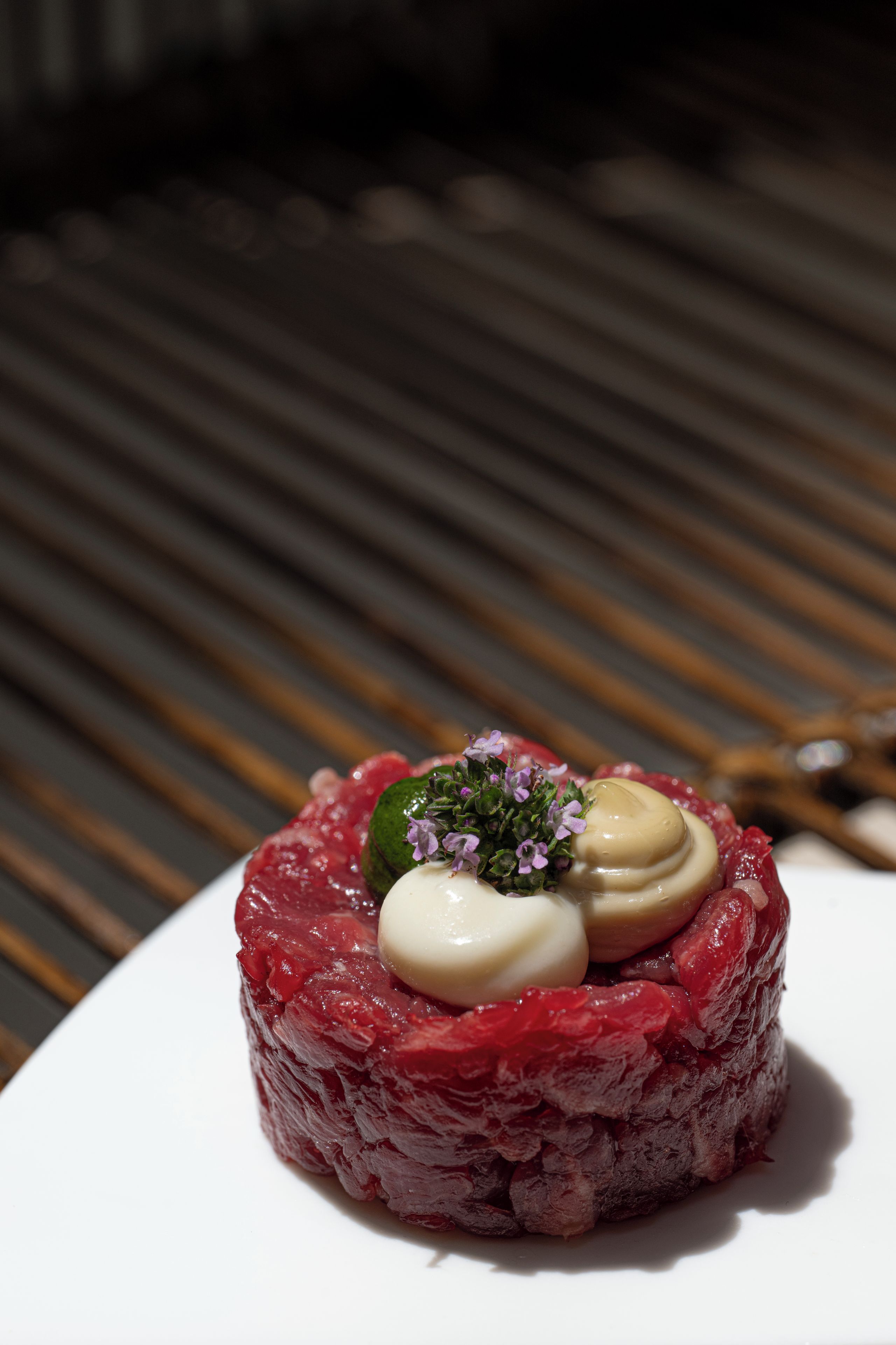 A dish with a round, layered structure, possibly a type of sushi or tartare, with a garnish of small purple flowers and a creamy topping.