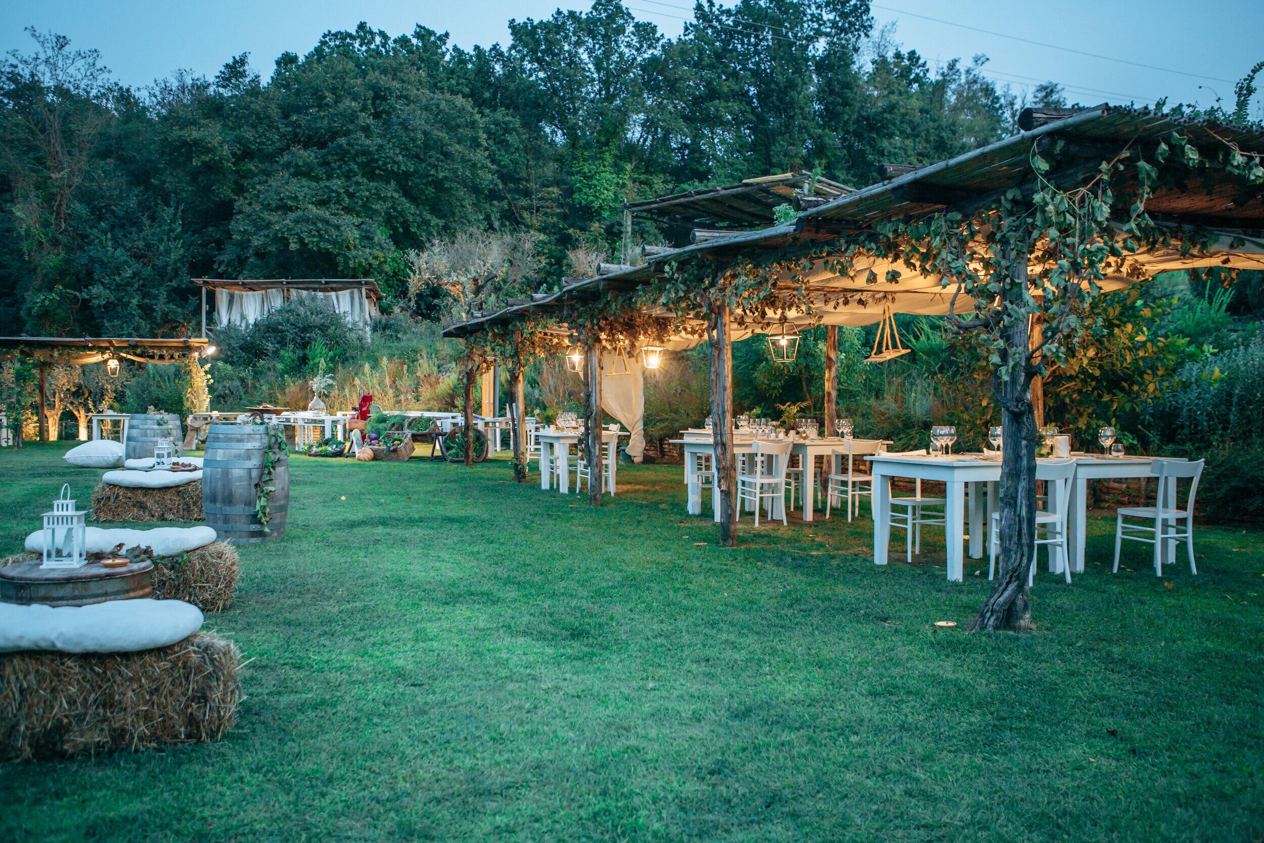 Un espacio de comedor al aire libre con mesas rústicas y fardos de heno, rodeado de follaje y una pérgola de madera con luces colgantes.
