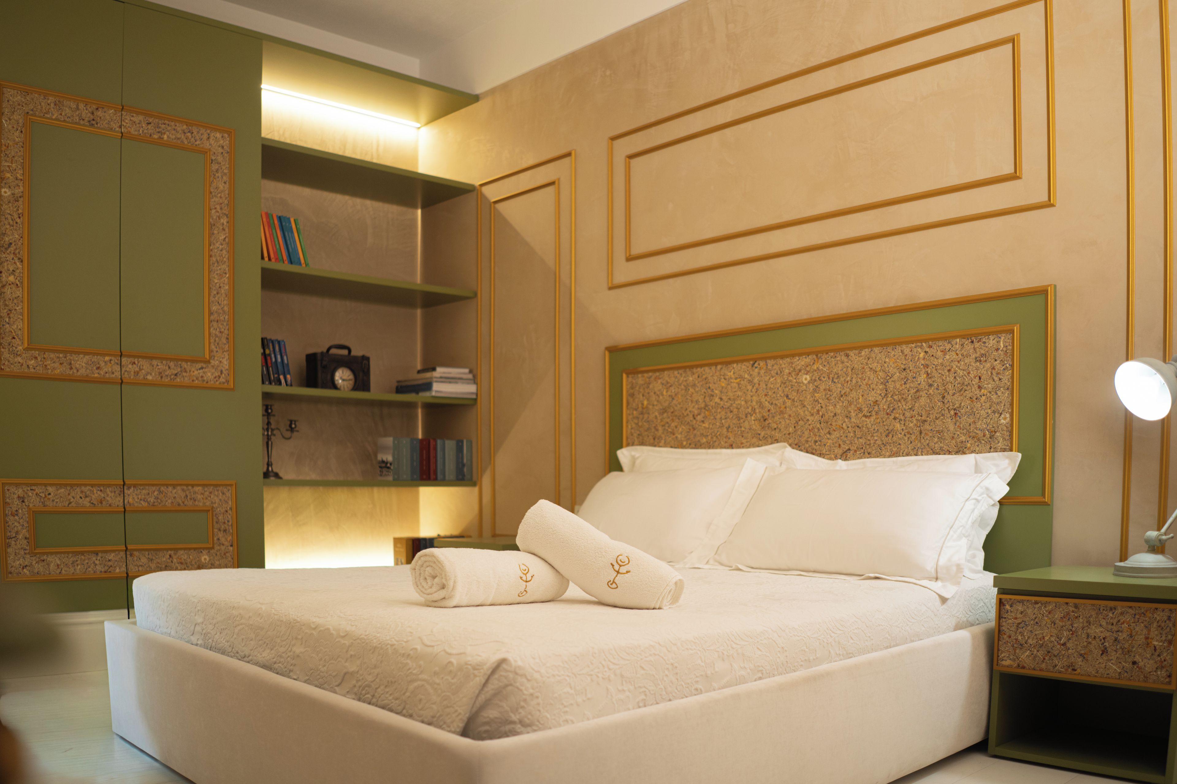 A neatly made bed with pillows and a folded towel, a bedside table with a lamp, and a bookshelf with books and a camera.