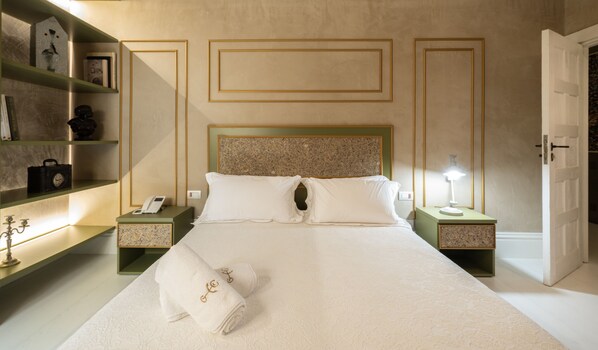 Habitación de hotel con una cama grande, dos mesitas de noche, una mesilla, un reloj de pared, un teléfono, una lámpara y una puerta.