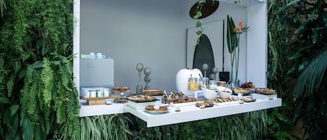 Une table de buffet extérieure blanche, garnie de divers aliments et condiments, et entourée d’un luxuriant feuillage verdoyant.