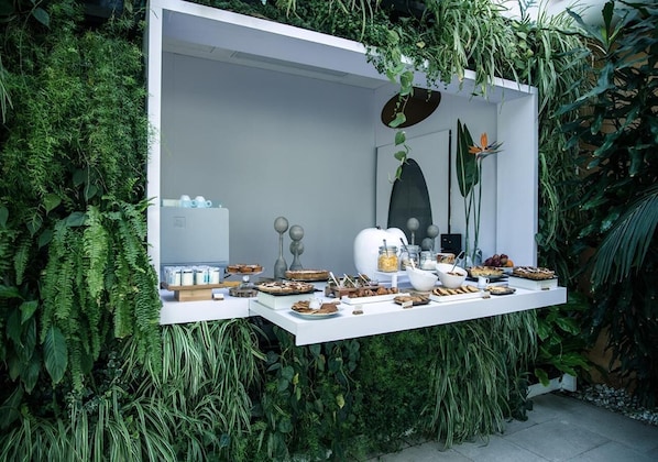 Una mesa buffet blanca al aire libre con diversos alimentos y condimentos, rodeada de abundante follaje verde.