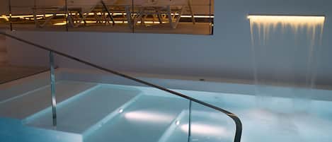 A modern indoor pool area with a glass barrier and a waterfall feature.