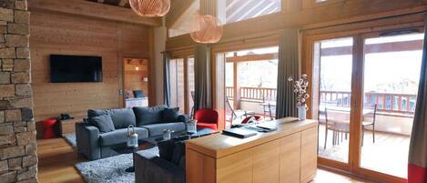 A modern interior with a wooden ceiling, a stone wall, a grey sofa, a wooden dining table, and a large window.