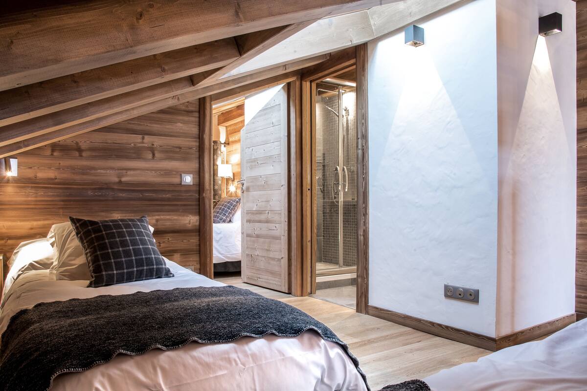 A bedroom with a sloped ceiling, wooden beams, a bed with a dark blanket, a nightstand, and a wall-mounted light.