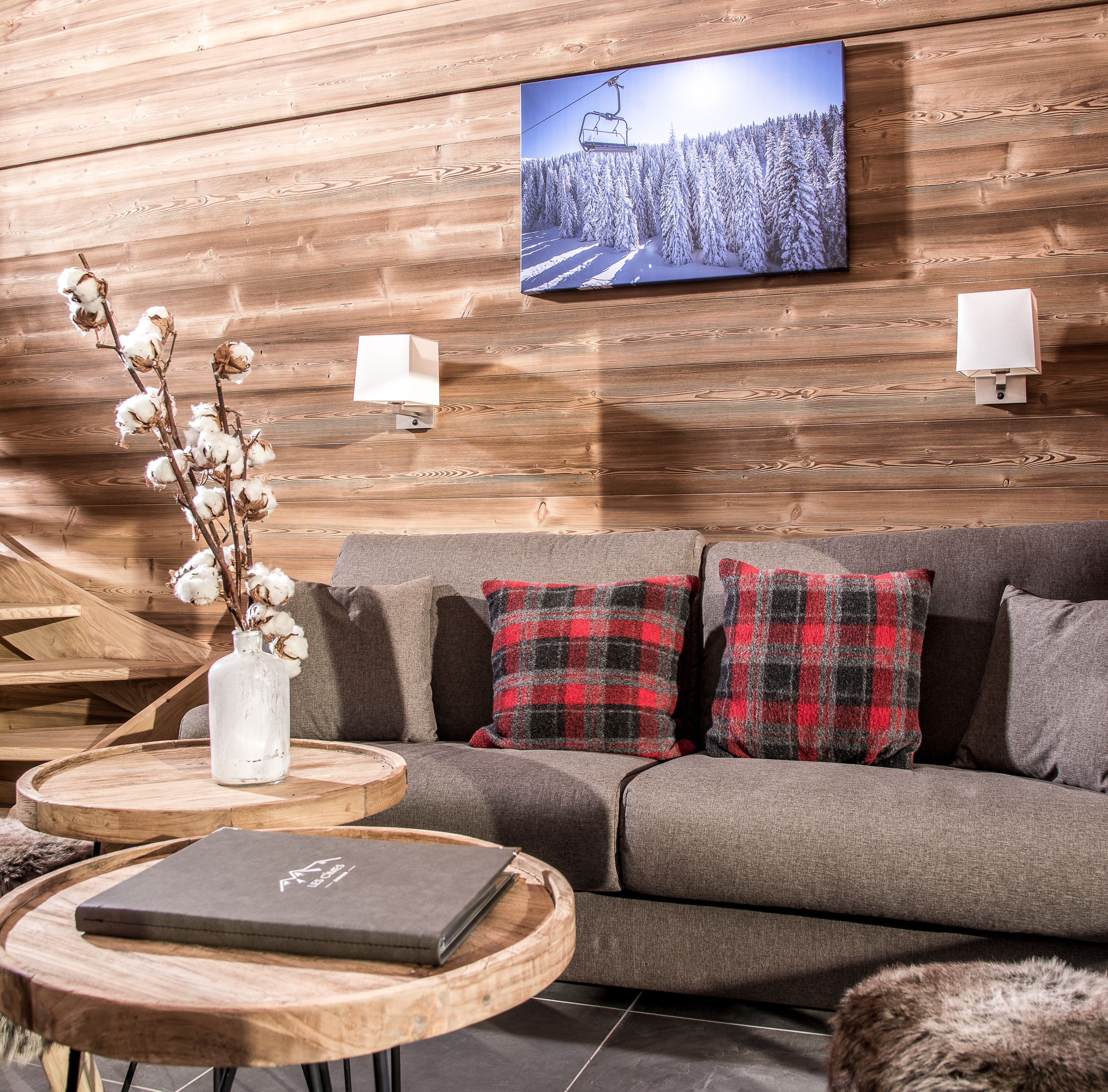 Une pièce chaleureuse avec un mur en bois, un canapé agrémenté de coussins à carreaux, un vase contenant des branches de coton, un ordinateur portable posé sur une table en bois et un téléviseur diffusant un paysage enneigé.
