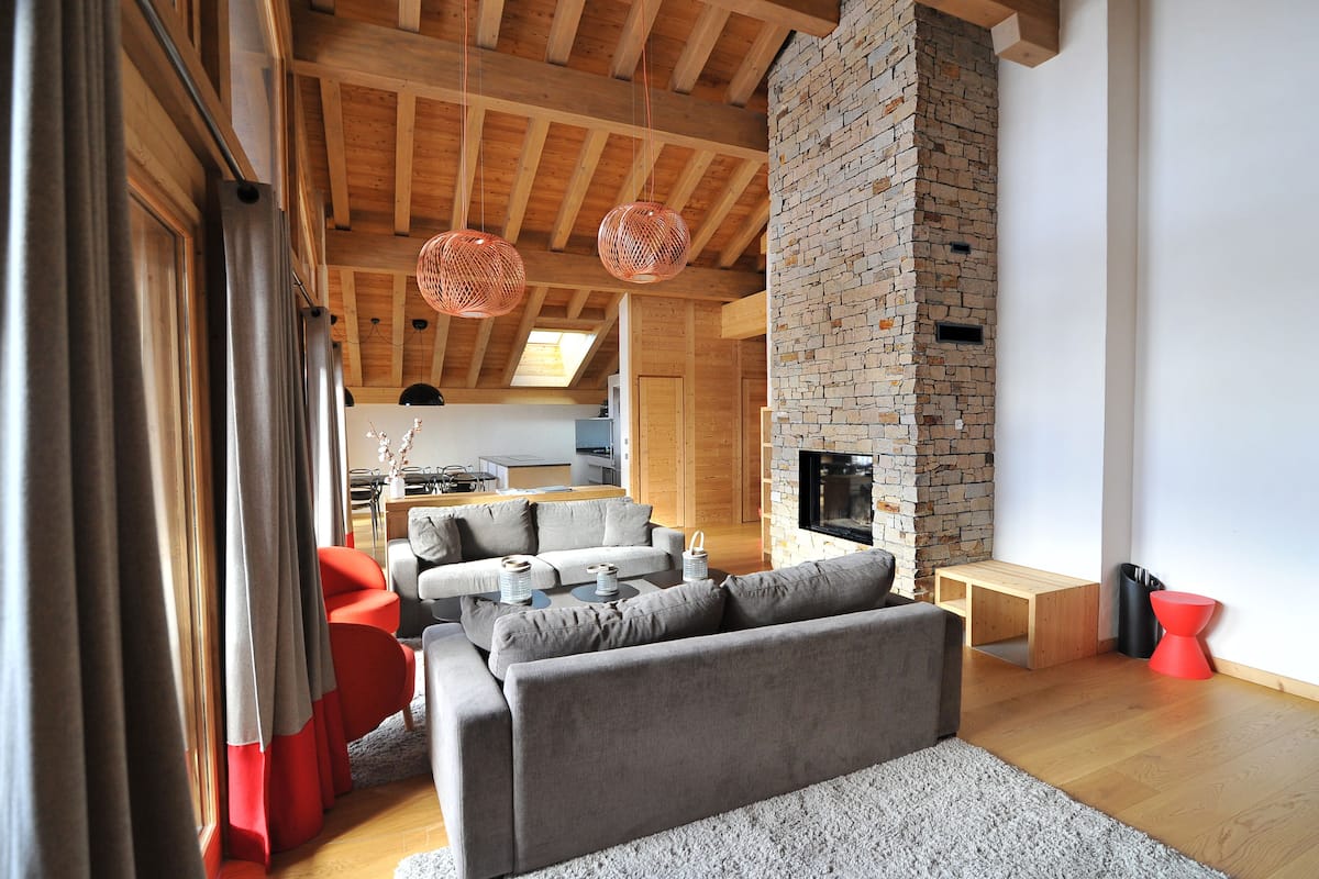 A modern living room with a grey sofa, red accent chairs, a fireplace, and wooden beams on the ceiling.