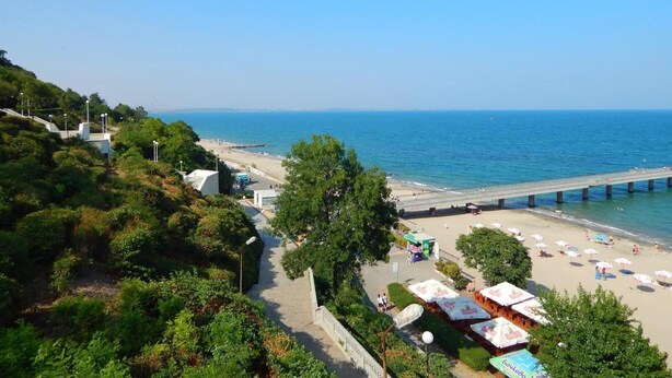 A coastal area with a beach, a pier, and a parasail in the sky. - Hotel Milano (Burgas)