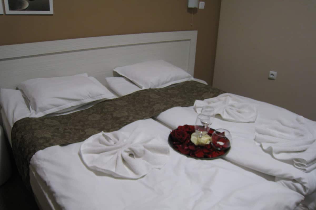 A hotel room with a neatly made bed, a tray with rose petals, a glass of wine, and towels.