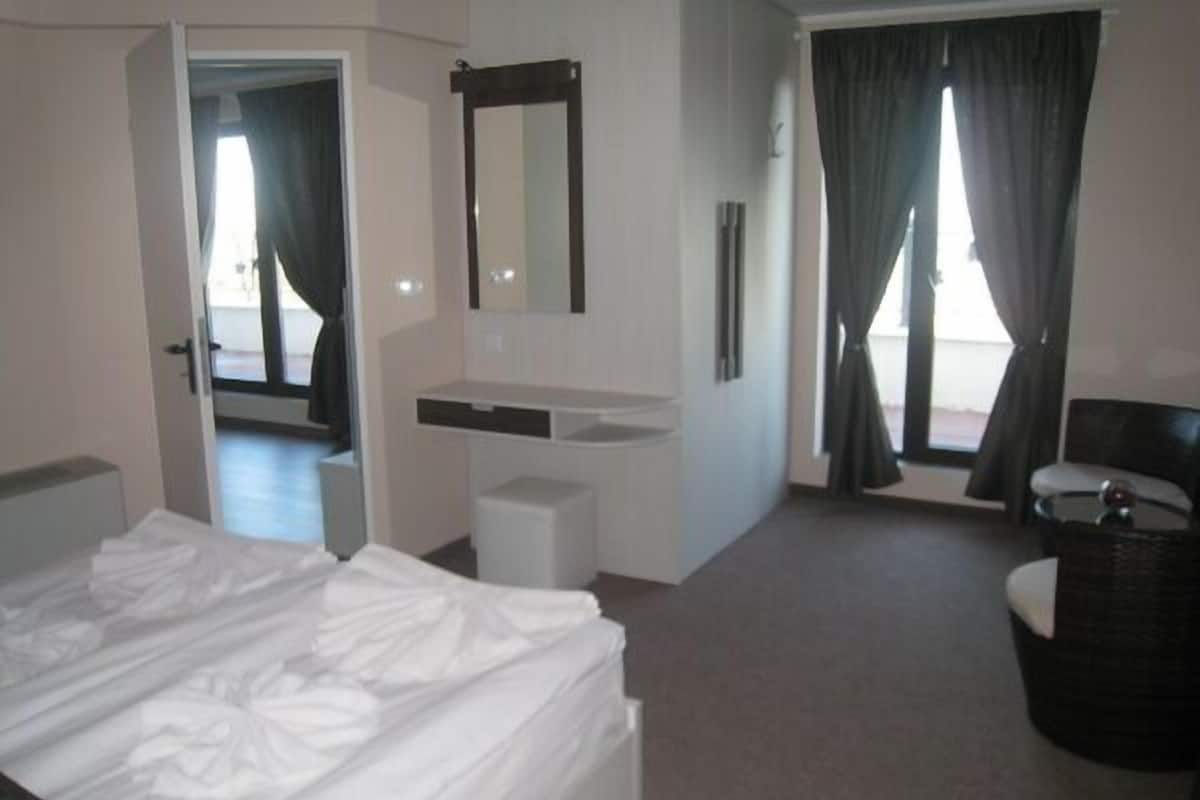 A hotel room with a bed, a small desk, a mirror, a window with curtains, and a wicker chair.