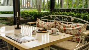A white metal bench with floral cushions, a wooden table with a white cup and saucer, a candle holder, and a menu. - Hotel Milano (Burgas)