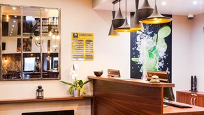 Un espace d’accueil d’hôtel comprenant un comptoir de réception en bois, un miroir, un tableau affichant le menu et une œuvre d’art murale représentant une tranche de citron vert.