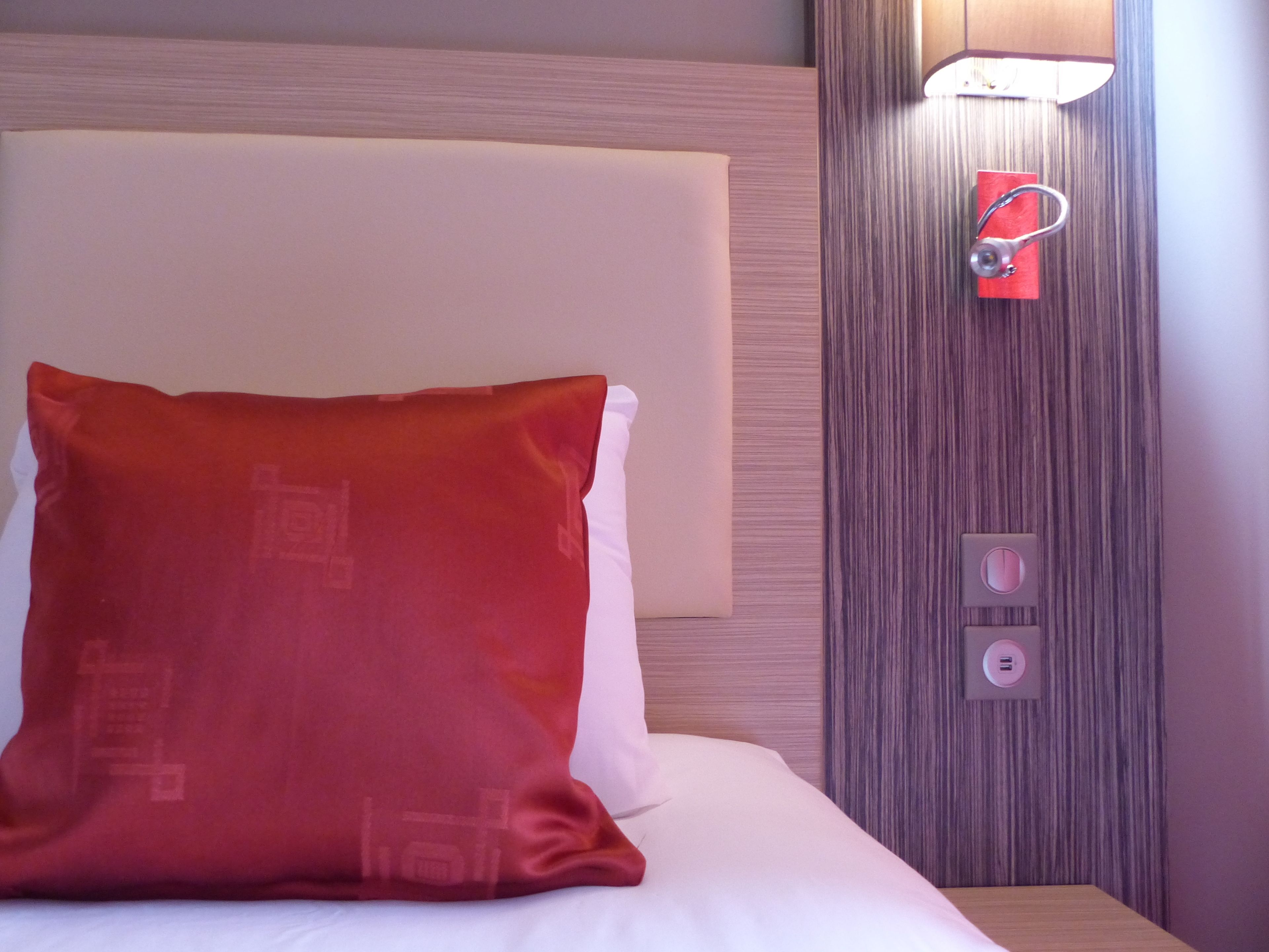 Ein Hotelzimmer mit einem Bett, einem roten Kissen, einer Wandleuchte und einem roten Gegenstand an der Wand.