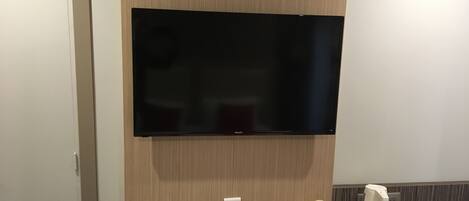 A hotel room with a flat-screen TV mounted on the wall, a small table with a remote control, a tray with snacks, and a coffee maker.