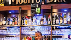 Un barman, derrière le comptoir d’un bar à mojitos, tient un shaker à cocktails. Le bar est doté d’étagères remplies de différentes bouteilles et de verres.