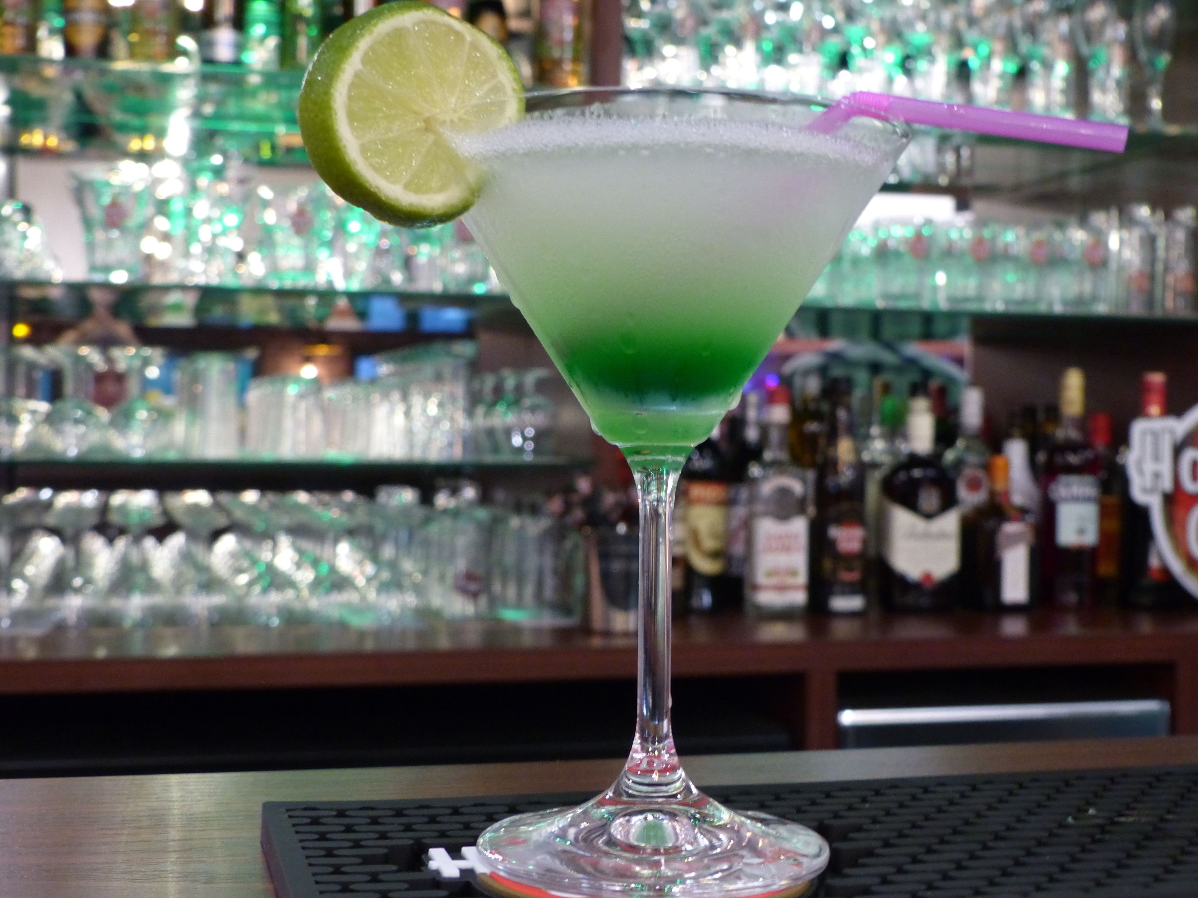 Un cocktail in un bicchiere da martini, guarnito con una fetta di lime, appoggiato su un bancone da bar con sullo sfondo scaffali pieni di bottiglie, sfocati.