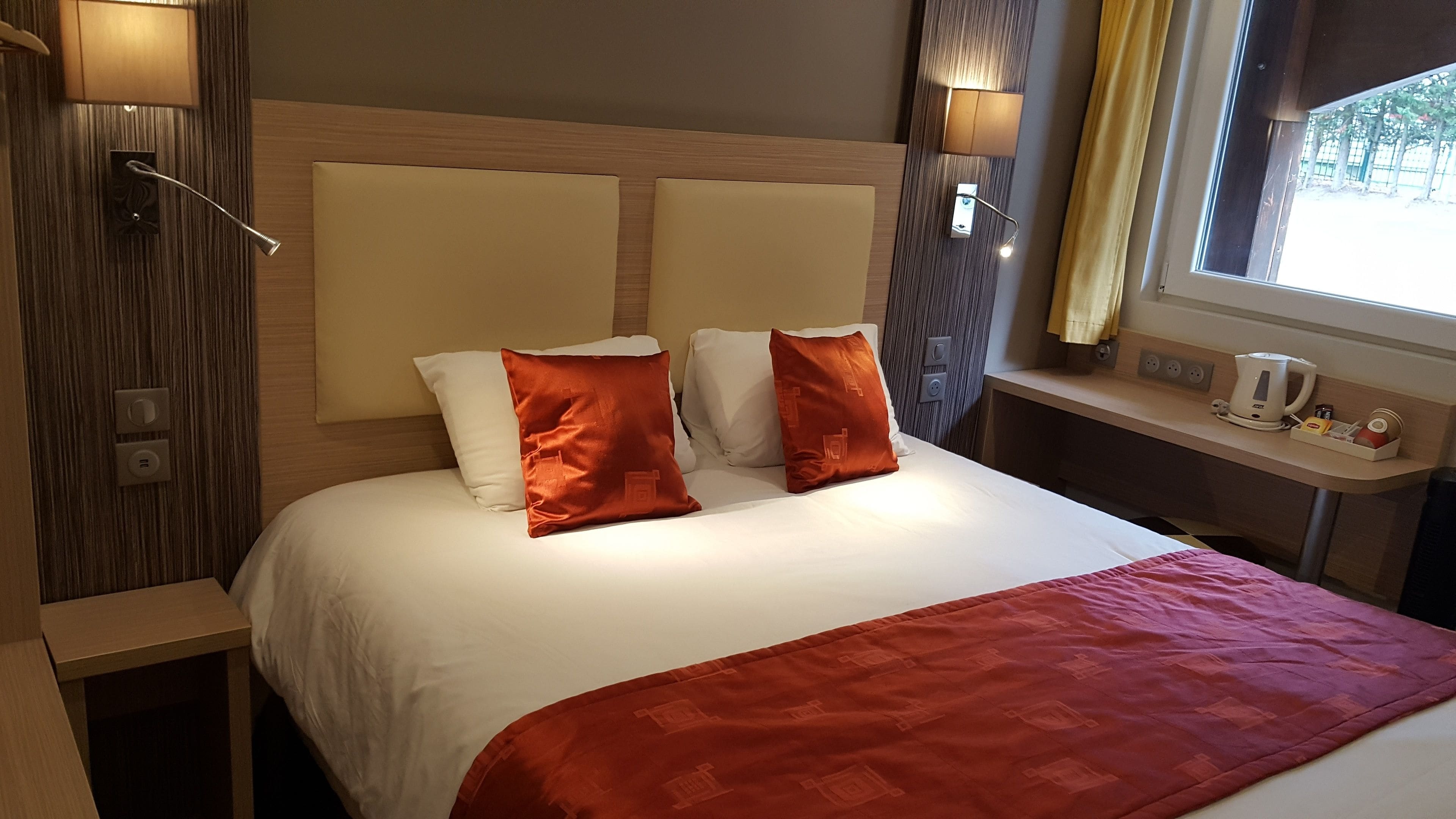 Habitación de hotel con una cama grande, dos lámparas de noche, un ventanal con cortinas, una mesita de noche con hervidor y un enchufe.