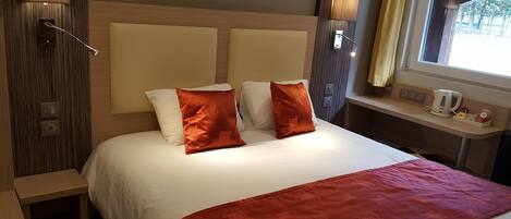 Habitación de hotel con una cama grande, dos lámparas de noche, un ventanal con cortinas, una mesita de noche con hervidor y un enchufe.
