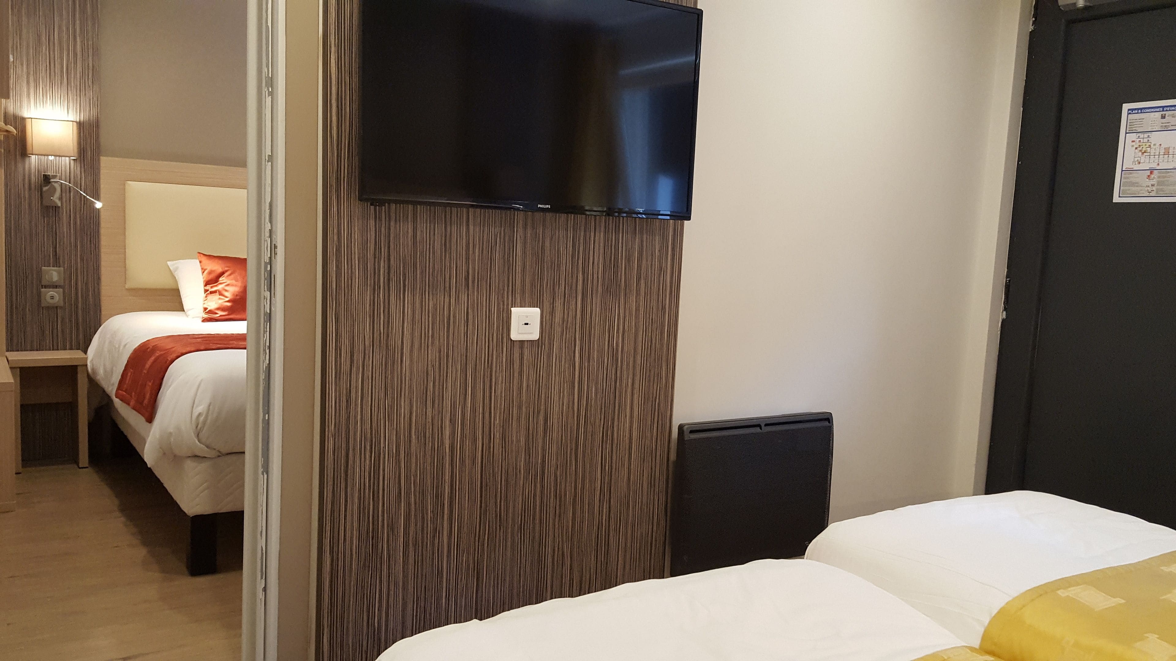Une chambre d’hôtel avec un lit, un téléviseur fixé sur un panneau en bois, une table de chevet et une lampe.