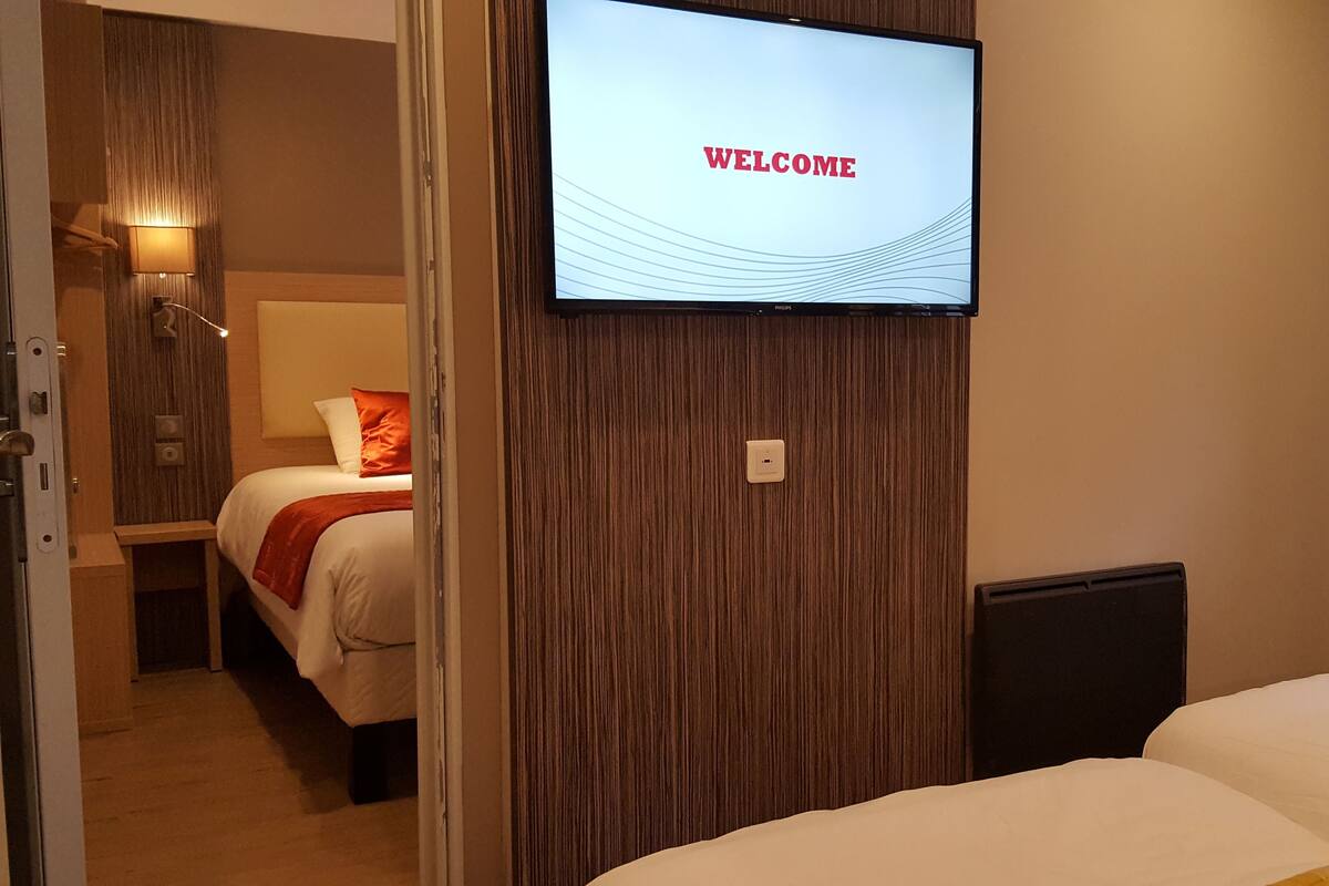 Habitación de hotel con una televisión mostrando la palabra “WELCOME”, una cama con ropa de cama blanca y amarilla, una pared de acento de madera y una mesita de noche con una lámpara.